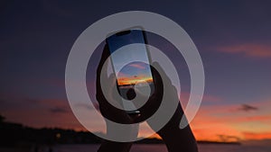 A girl photographs a colorful tropical sunset on a modern close-up phone. Silhouette of a woman's hands taking a