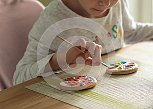 Girl with pigtails paints Easter glazed cookies with a brush while sitting in the kitchen at the table