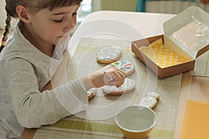 Girl with pigtails paints Easter glazed cookies with a brush while sitting in the kitchen at the table