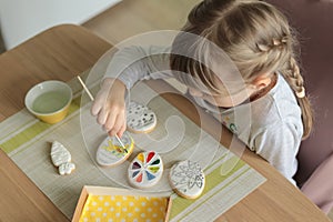 Girl with pigtails paints Easter glazed cookies with a brush while sitting in the kitchen at the table