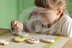 Girl with pigtails paints Easter glazed cookies with a brush while sitting in the kitchen at the table