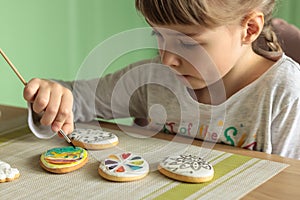 Girl with pigtails paints Easter glazed cookies with a brush while sitting in the kitchen at the table