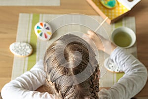 Girl with pigtails paints Easter glazed cookies with a brush while sitting in the kitchen at the table