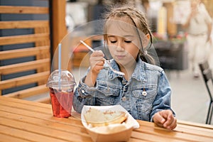 A girl with pigtails sits at a table in the courtyard of a restaurant and eats with the pleasure of takeaway food.