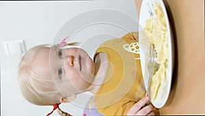 A girl with pigtails is sitting at the table and eating Farfalle pasta with a fork.