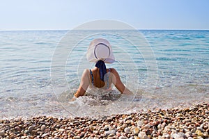 Girl in pink bikini with white hat sitting back to the camera on pebbles on the beach, sunbathing and looking at pure