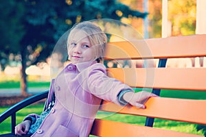 A girl in a pink coat is sitting on a yellow bench in the park.