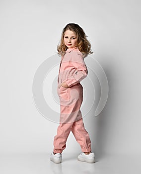 girl in a pink cotton jumpsuit stands with her back to the camera and turned with her legs wide apart, her hand in her