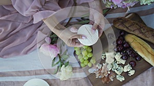 A girl in a pink dress prepares a meal at a picnic in the summer. Cheese, grapes, cherries, nuts, flowers.