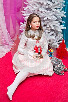 A girl in a pink elegant dress with Santa Claus in her hands sits on a red armchair by the Christmas tree