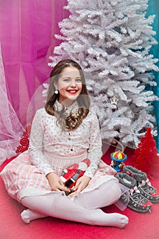 A girl in a pink elegant dress with Santa Claus in her hands sits on a red armchair by the Christmas tree