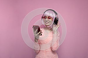 Girl with pink hair in headphones listens to music and holds a smartphone, hipster uses a phone on a pink background