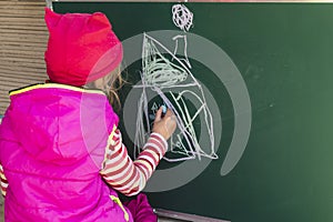 A girl in a pink jacket draws with chalk on a school blackboard a house. In her hands is chalk