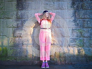 A girl in a pink jumpsuit stands holding her hands behind her head near a stone wall in the sunlight