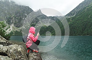 Girl in a pink raincoat sits on a rocky stone against the background of a large mountain lake in the rain and takes a photo on a