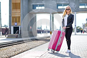 A girl with a pink suitcase is standing at the railway station