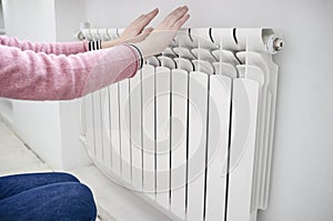 Girl in a pink sweater and blue jeans warms hands near a radiator