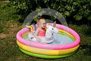 Girl in a pink swimsuit and sunglasses swims in a small colorful inflatable pool in the garden of the house on a hot summer sunny