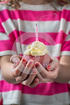 A girl in a pink and white shirt holding a pink cupcake in two hands with candle just blown out, vertical