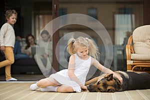 Girl playing with dog outside, family inside house at background