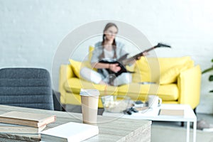 girl playing electric guitar on sofa boks and disposable cup of coffee on table