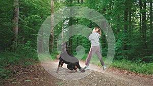 Girl playing with her jumping rottweiler dog with a ball in a green forest on a path in the sunlight