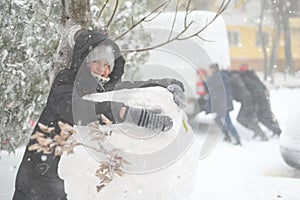 a girl is playing with snow on the street, making a snowman, on background a car is stuck in snow and men are pushing, it is