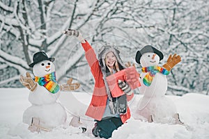 Girl playing with snowman in winter park. Snowman and funny young woman in winter hat and scarf.