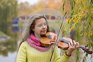 Girl playing the violin eyes closed in the autumn park at a lake and willow foliage background