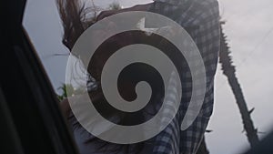 Girl with pleasant smile adjusts hair leaning out of car window traveling along road with palm trees