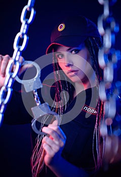 A girl in a police uniform with dreadlocks in neon light with chains and handcuffs English translation the police