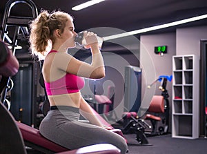 Girl posing in the gym with a bottle in her hands, sitting on a bench. Horizontal photo