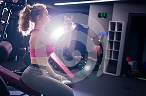 Girl posing in the gym with a bottle in her hands, sitting on a bench. Horizontal photo