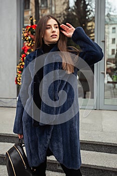 Girl posing on road on winter background. Glamorous funny young woman with smile wearing stylish blue long fur coat