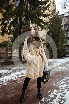 Girl posing on road on winter background. Glamorous funny young woman with smile wearing stylish creamy long fur coat