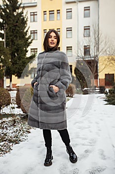 Girl posing on road on winter background. Glamorous funny young woman with smile wearing stylish grey long fur coat