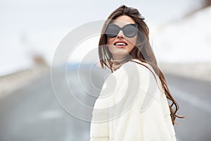 Girl posing on road on winter woods background. Glamorous funny young woman with smile wearing stylish sweater, white