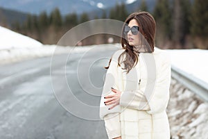 Girl posing on road on winter woods background. Glamorous funny young woman with smile wearing stylish sweater, white