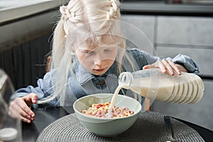 Girl pouring milk from the bottle to the plate while sitting at the kitchen