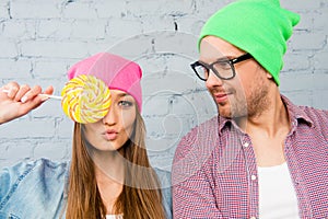 Girl pouting and holding candy near eyes with her hipster boyfriend in glasses and cap