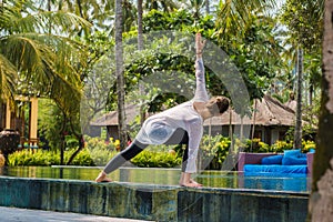 Girl is practicing yoga on the edge of swimming pool with green plants and palm trees around, stretching legs