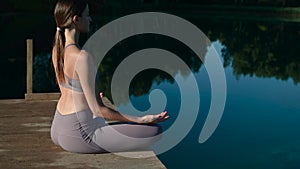 Girl is practicing yoga technique. She is sitting on the pier in lotus pose by the lake and meditating. Young slim woman