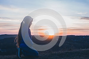 The girl is praying on her knees. On the background of the sunset sky. Repentance for sins. Kneeling Prayer to God. Worship