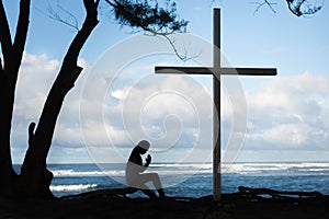 Girl praying to God in front of a cross with a beautiful blue ocean background.