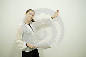 Girl is presenting something while standing by a white wall holding sheets in one hand and showing with the other hand