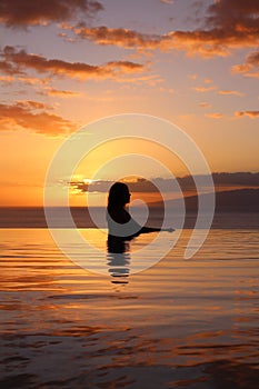 Girl in profile stands in the pool, against the background of an orange sunset and the ocean. hotel