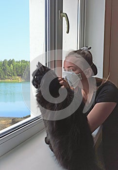 Girl in protective mask with cat looking sadly out the window during quarantine