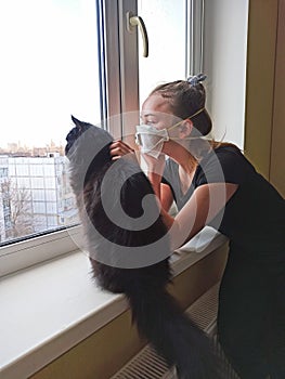 Girl in protective mask with cat looking sadly out the window during quarantine