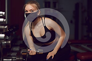 The girl in a protective mask is engaged in dumbbells. Protective masks against virus infection. Training during quarantine in the