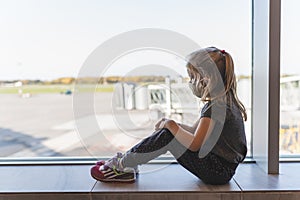 Girl with protective mask on her face look sad through the window at airport on planes. Travel, pandemy